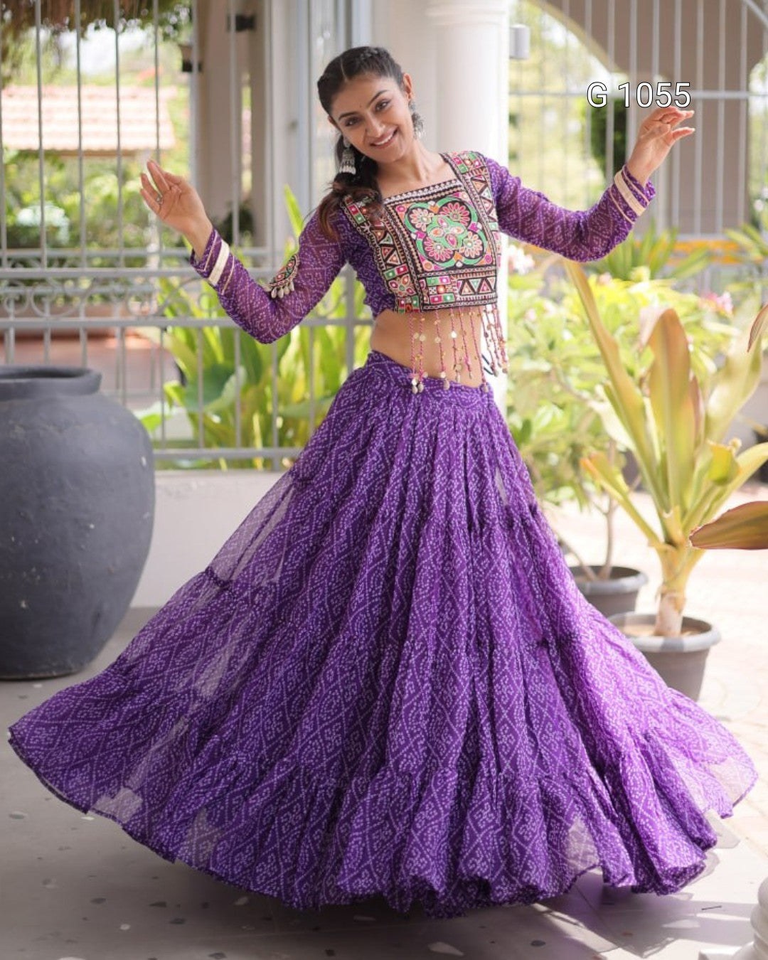 A woman modeling a purple embroidered traditional Indian lehenga choli set, featuring a long-sleeved embroidered top and a full flared skirt, posed outdoors on a patio.