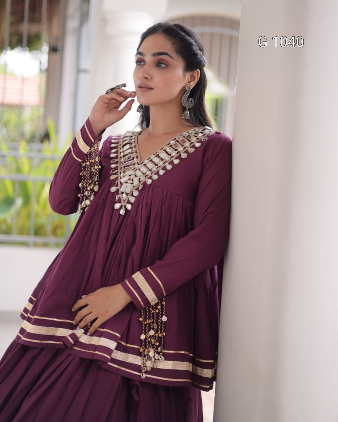 A woman wearing a deep purple traditional kurta set with intricate silver and gold embroidery, standing next to a white pillar in a bright, modern indoor setting.
