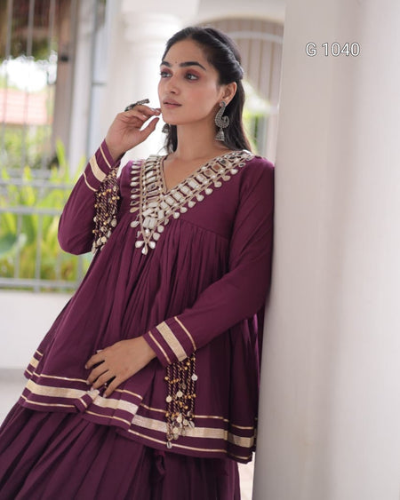 A woman wearing a deep purple traditional kurta set with intricate silver and gold embroidery, standing next to a white pillar in a bright, modern indoor setting.