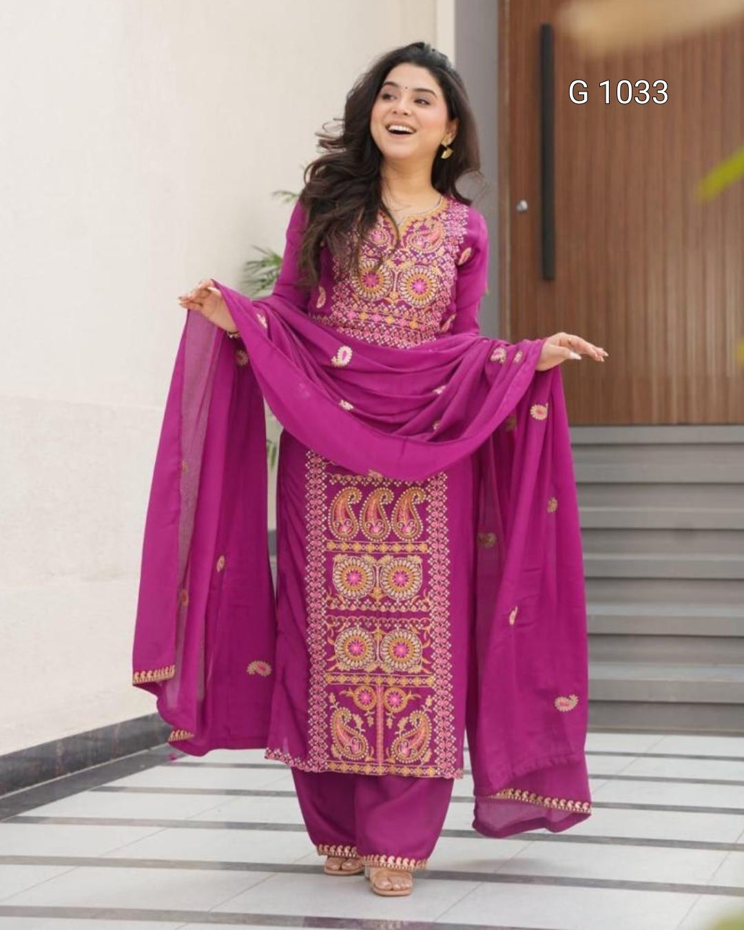 A woman modeling a magenta pink salwar kameez suit with intricate gold and yellow embroidery, holding out a matching sheer dupatta in front of a modern building entrance.