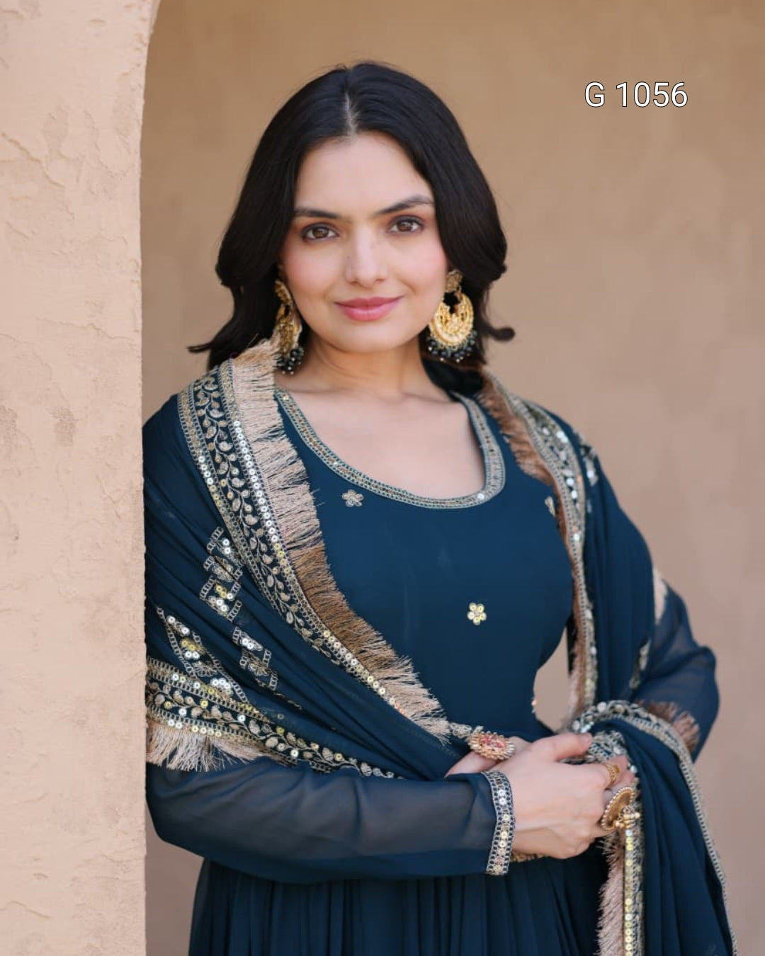 A woman wearing a dark teal blue traditional outfit with gold and silver embroidery and a matching dupatta, standing against a beige textured wall. The text 'G 1056' is visible in the top right corner.