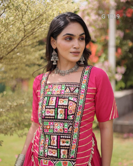 A woman wearing a bright pink traditional kurta with colorful geometric embroidery and mirror work on the front panel, photographed outdoors with blurred greenery in the background.