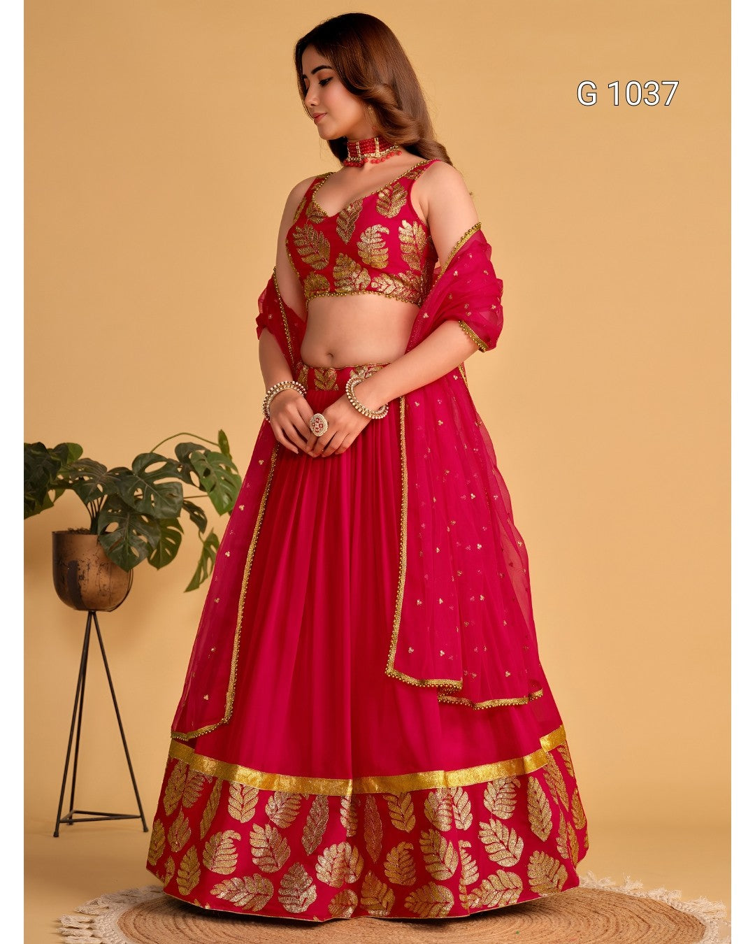 A woman models a bright pink and gold traditional Indian lehenga choli set against a solid beige background. The outfit features a heavily embroidered gold leaf-patterned pink blouse and a matching flared skirt with a decorative gold border.