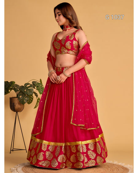 A woman models a bright pink and gold traditional Indian lehenga choli set against a solid beige background. The outfit features a heavily embroidered gold leaf-patterned pink blouse and a matching flared skirt with a decorative gold border.