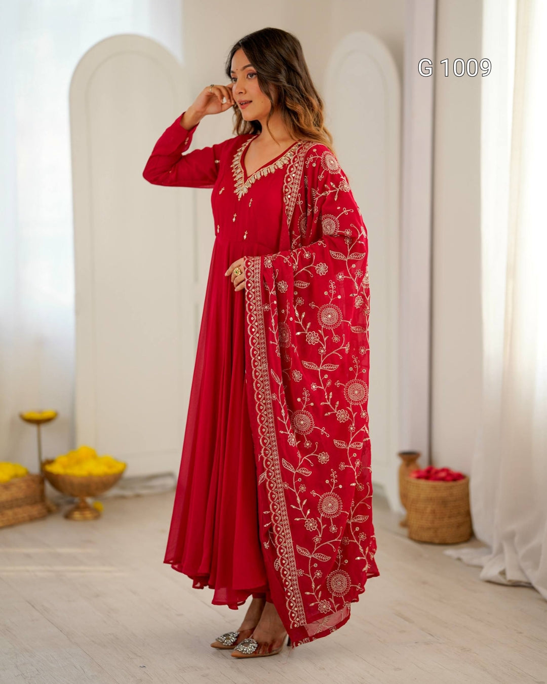 A woman models a traditional red long kurta with gold embroidery and a matching printed dupatta in a bright, white-walled room with arches.