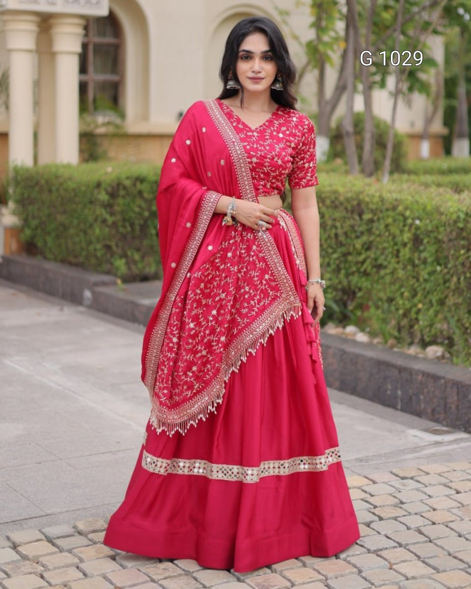 A woman modeling a bright pink-red lehenga choli set with gold embroidery, standing outdoors in front of a building and hedges. The set includes a heavily embroidered blouse, flared skirt, and draped dupatta.