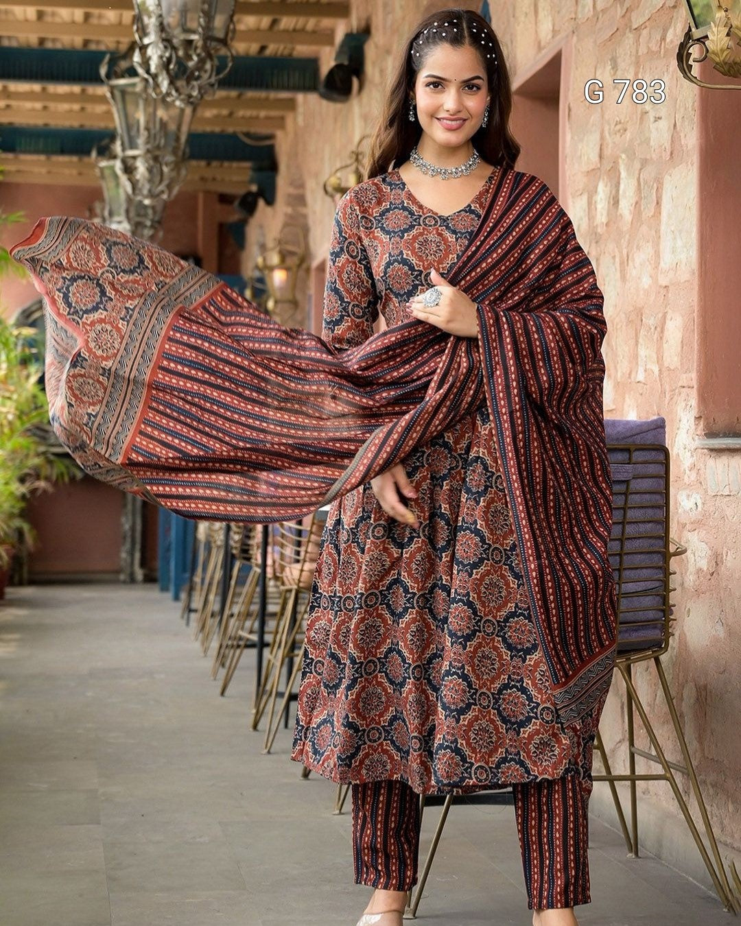 Woman in a patterned outfit standing in an outdoor setting with chairs and plants.