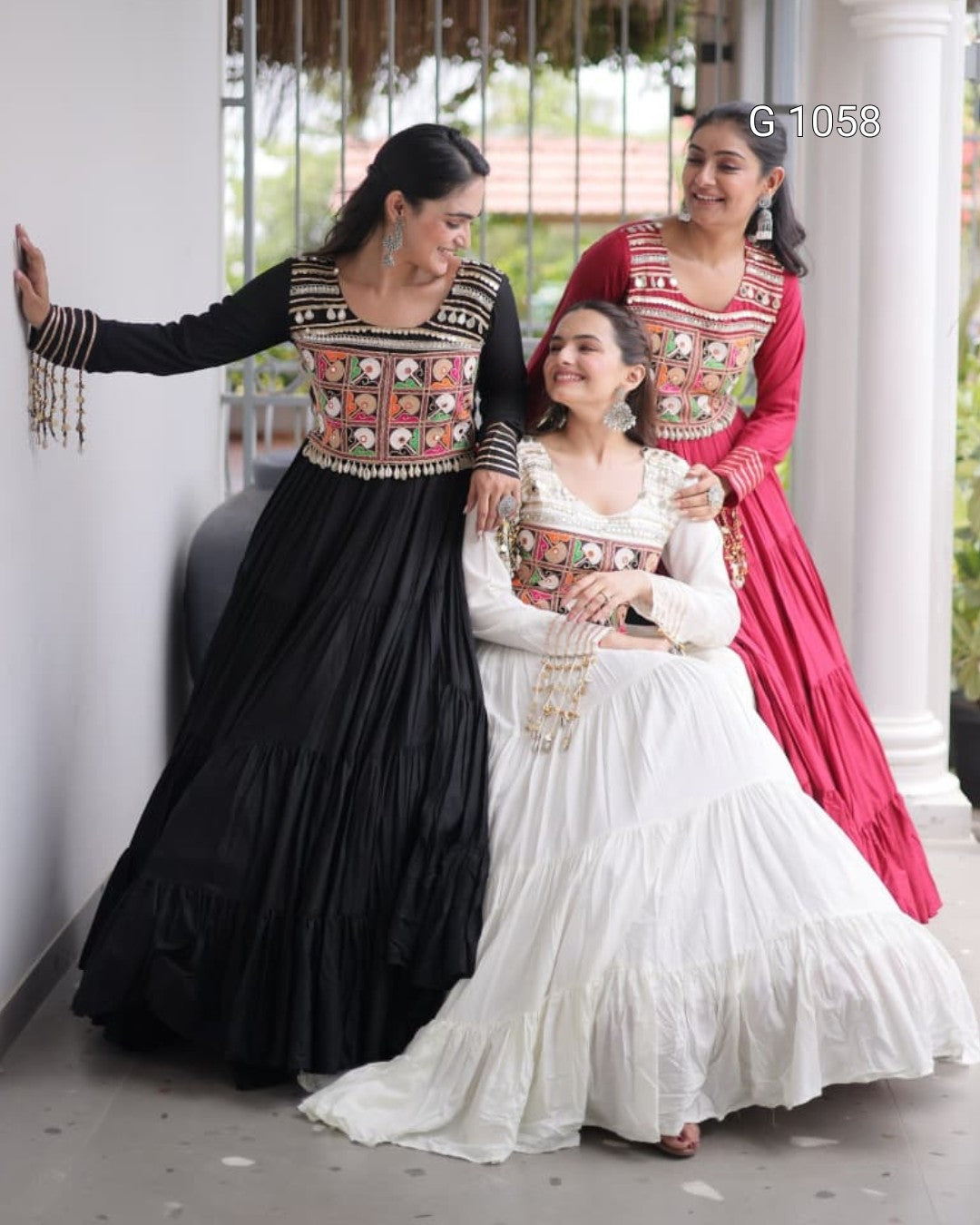 Three women wearing traditional Indian ethnic dresses with intricate embroidery, standing together on a porch. The dresses are in black, white, and red, featuring heavily embellished borders.