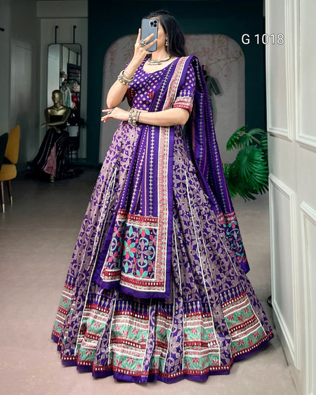A woman takes a mirror selfie wearing a purple Indian ethnic lehenga choli set with intricate multicolor embroidery and patterns. The set features a purple flared skirt (lehenga), a matching embroidered blouse, and a coordinating dupatta.