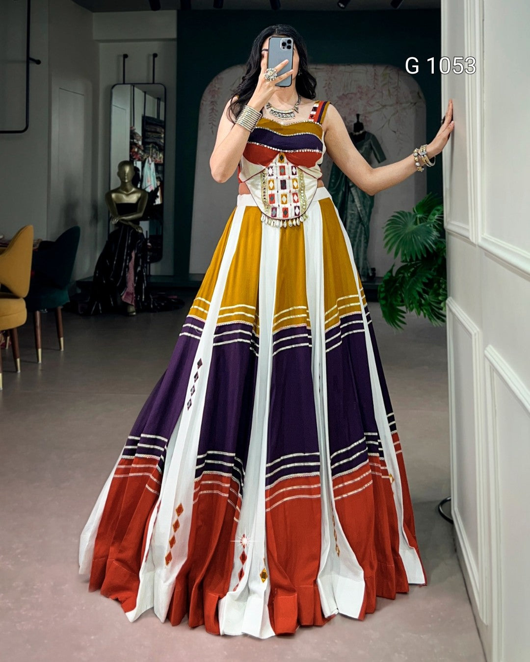A woman takes a mirror selfie in a boutique wearing a sleeveless, floor-length A-line gown with a geometric color-blocked pattern in mustard yellow, white, purple, rust red, and black.