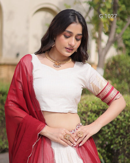 A woman wearing a white sleeveless crop blouse with red patterned cuffs, a white skirt, and a red dupatta, standing outdoors with a blurred garden background. The text 'G 1027' is visible in the top right corner.