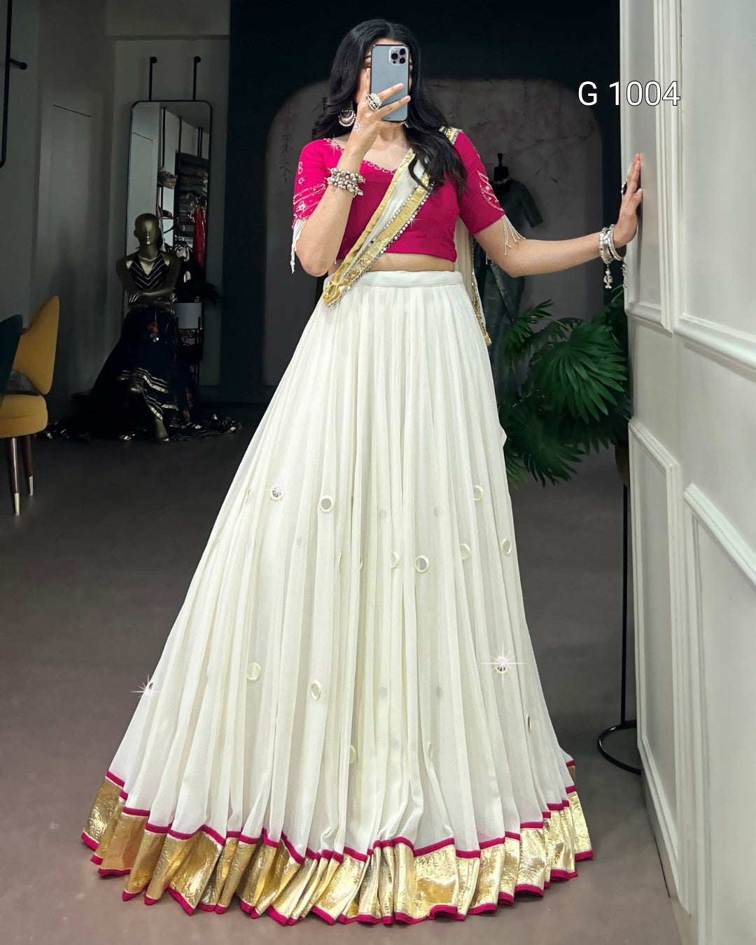 A woman takes a mirror selfie wearing a traditional Indian outfit consisting of a white flared skirt with gold and pink borders, a bright pink blouse with gold embroidery, and a gold and pink draped sash. The background shows a boutique interior with a mannequin and plants.