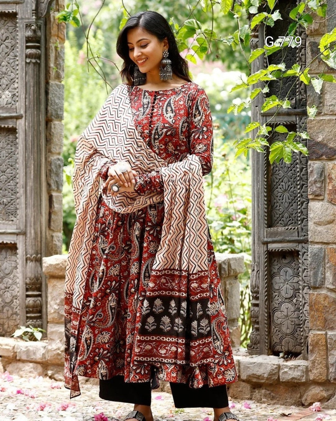 Woman in a patterned outfit standing in a garden with stone architecture.