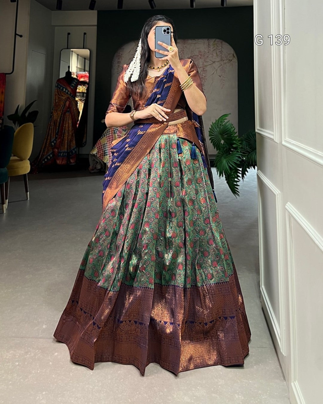 Woman taking a mirror selfie wearing a traditional saree in a room with white walls and a yellow chair.