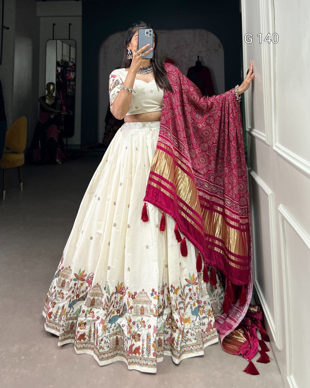 Woman in a white traditional outfit with a pink and gold dupatta, standing in a room.