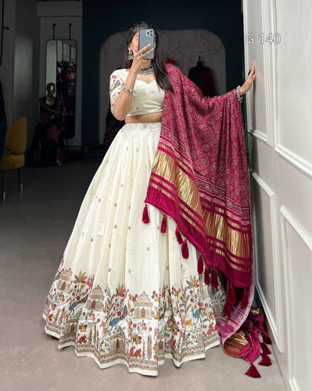 Woman in a white traditional outfit with a pink and gold dupatta, standing in a room.