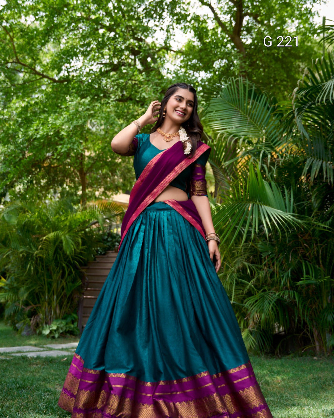Woman in a teal and purple saree standing outdoors with greenery in the background