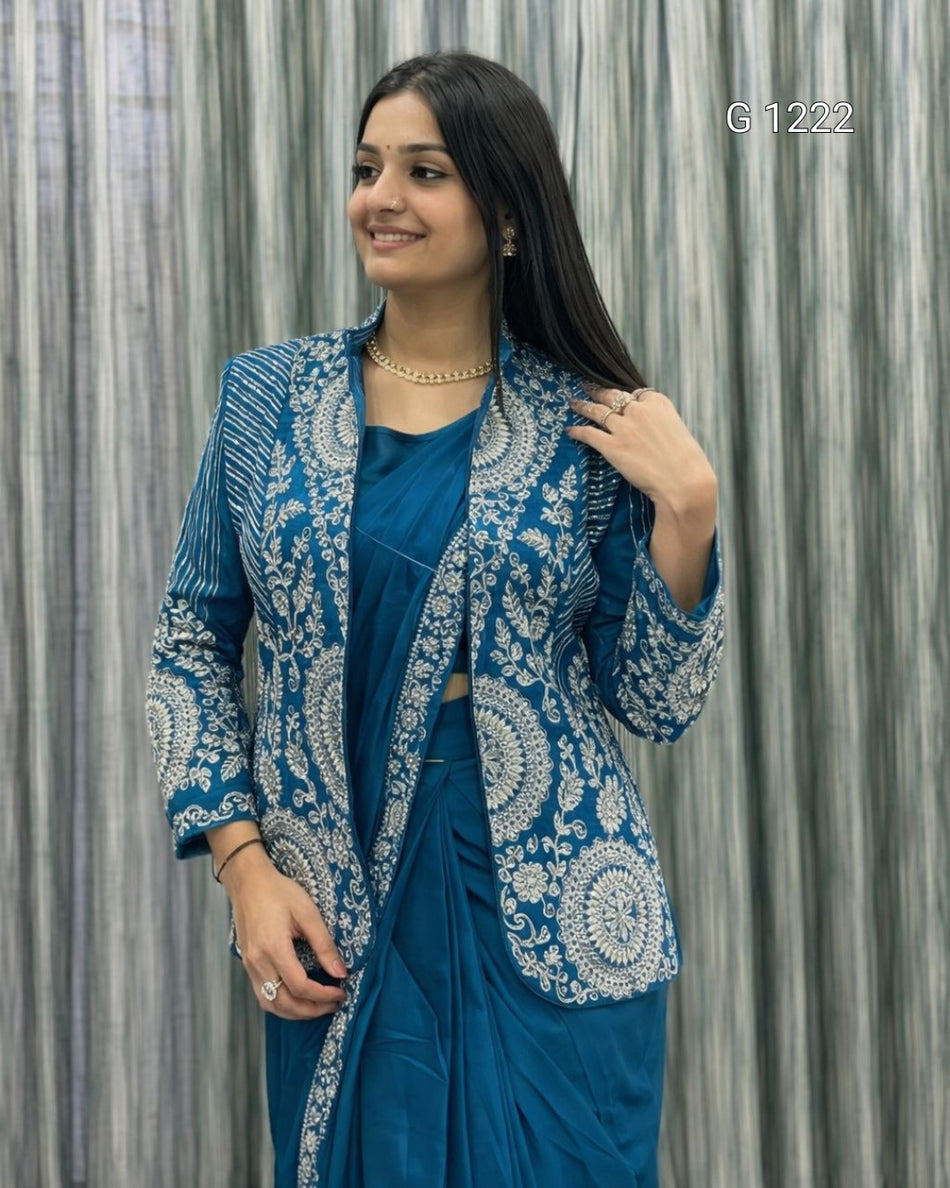 A woman modeling a teal blue traditional outfit with intricate white embroidery, consisting of a kurta, sari, and matching jacket, set against a gray textured curtain backdrop.
