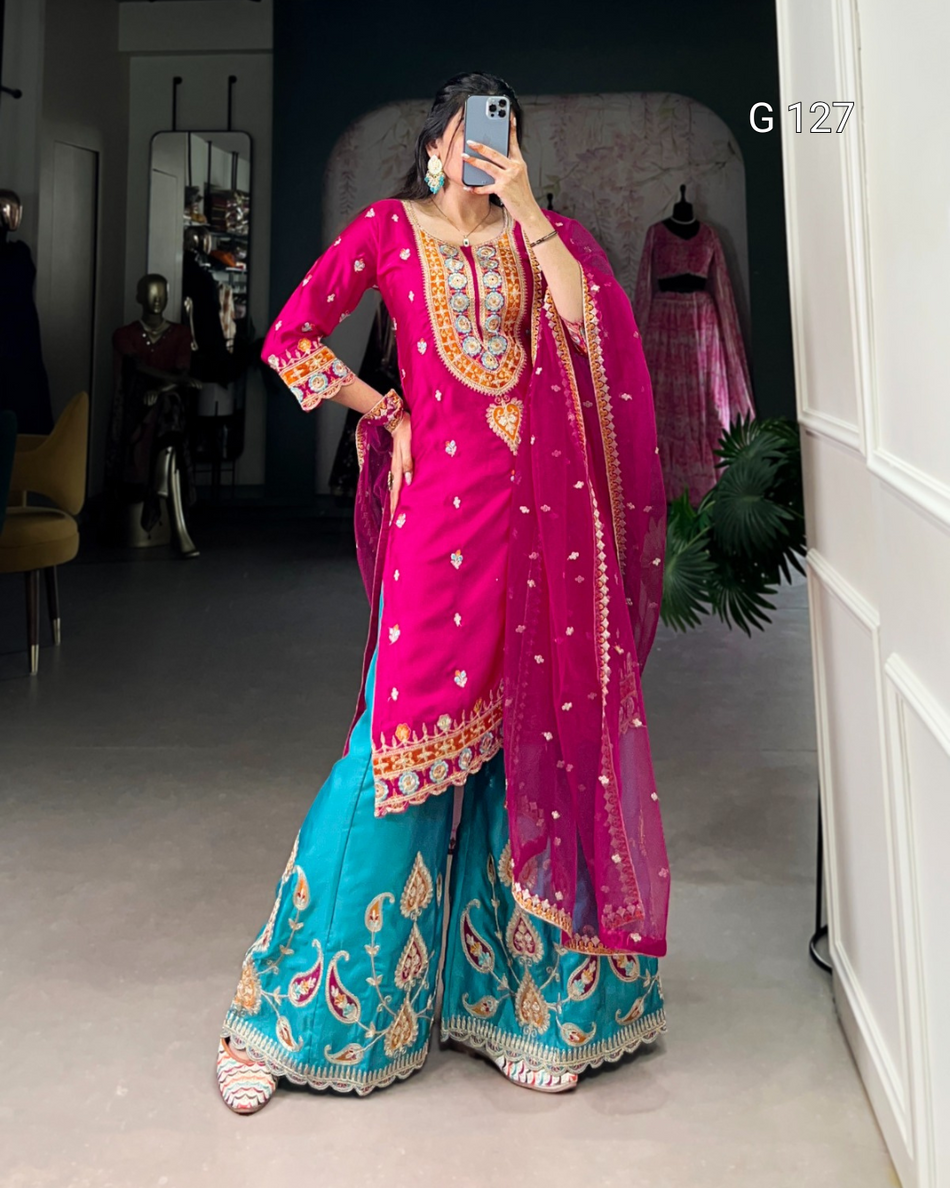 Person wearing a vibrant pink and blue traditional outfit with intricate designs, standing indoors.