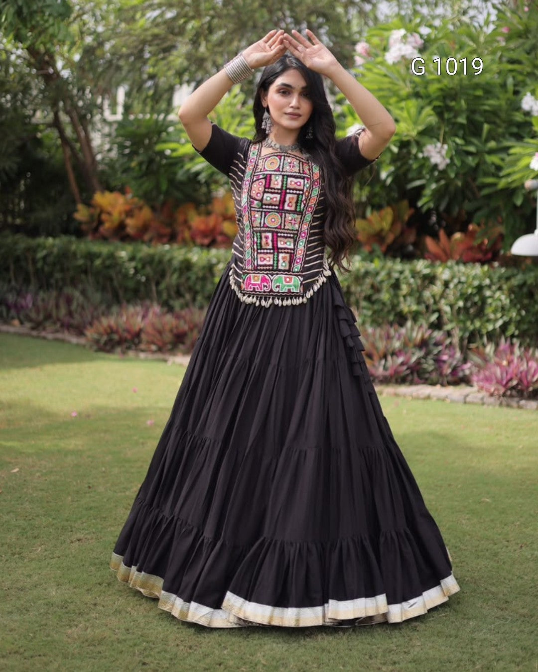 A woman stands outdoors on a grassy lawn wearing a black traditional long dress with a heavily embroidered multicolor bodice and a tiered skirt with a beige border. The background shows green hedges and trees.