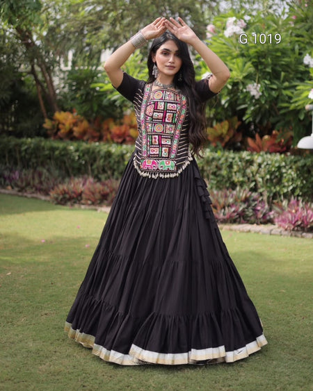 A woman stands outdoors on a grassy lawn wearing a black traditional long dress with a heavily embroidered multicolor bodice and a tiered skirt with a beige border. The background shows green hedges and trees.