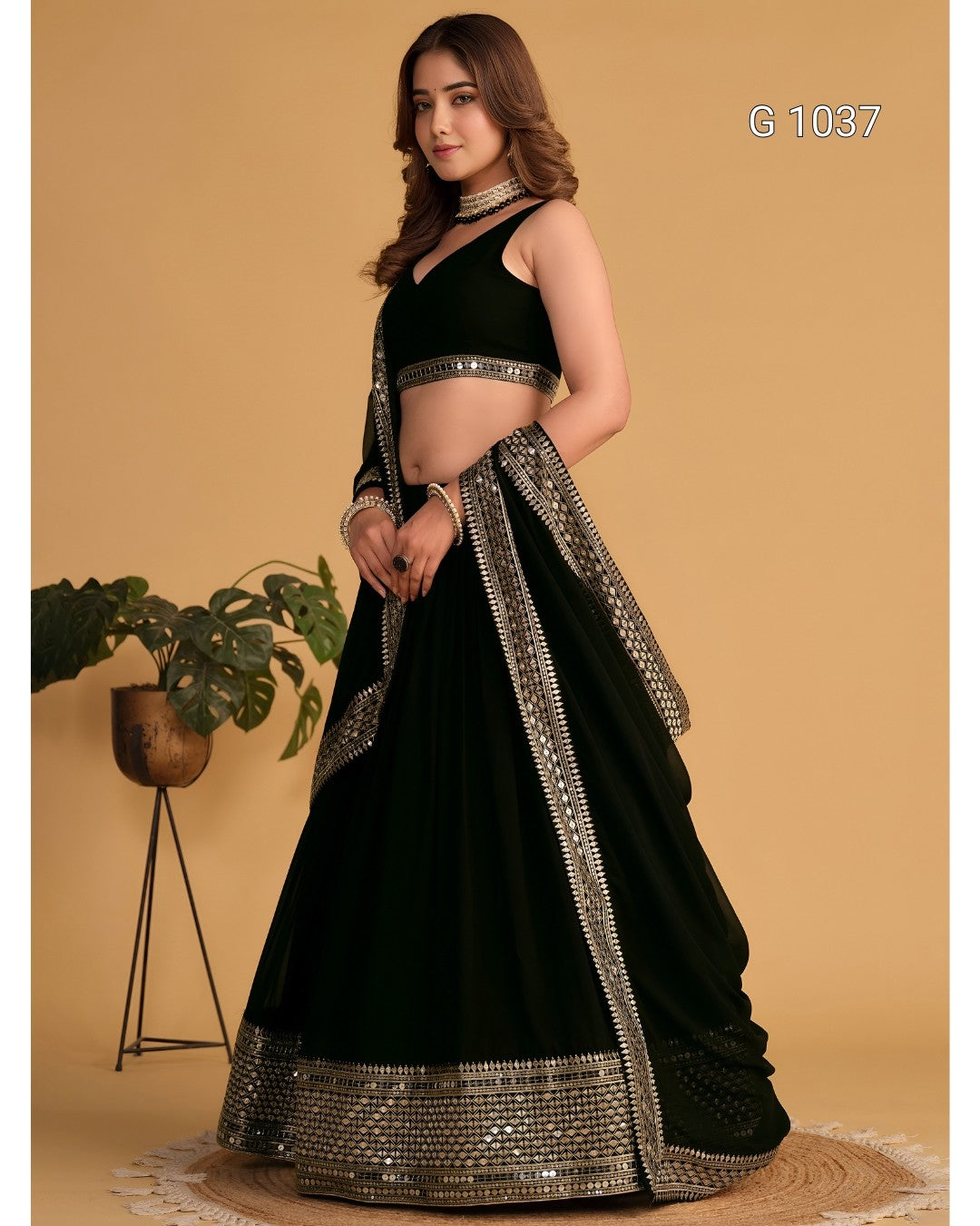 A woman models a black traditional lehenga choli set with ornate silver mirror-work borders, set against a warm beige background with a small potted plant on the left.