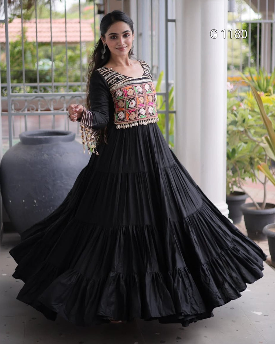 A woman modeling a black tiered maxi dress with a heavily embroidered multicolor bodice, standing in a covered outdoor walkway with potted plants and columns.