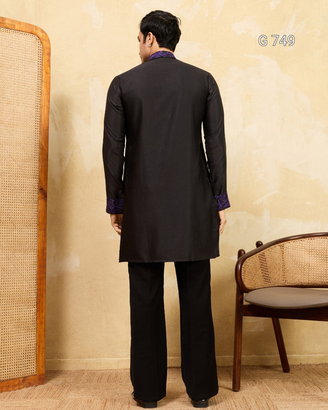 Man wearing a black traditional outfit standing against a beige wall with a chair and mirror in the background.