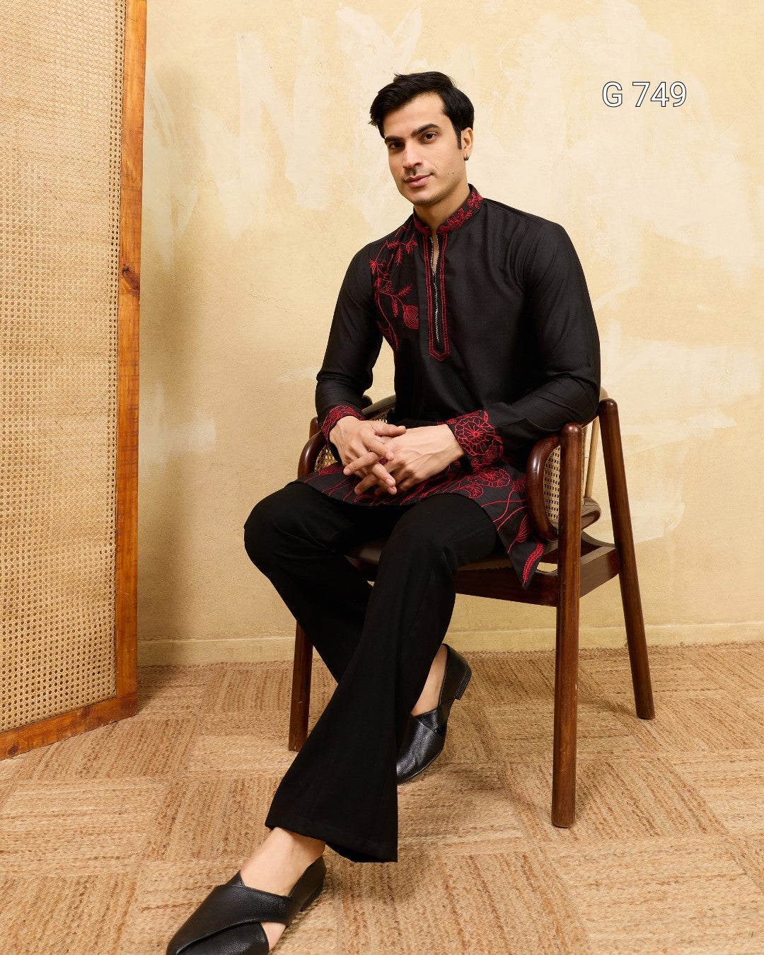 Man sitting on a wooden chair wearing a black kurta with red patterns against a beige wall.