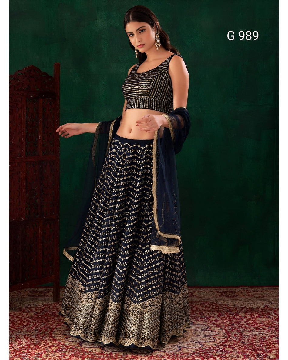 A woman models a black and gold embroidered Indian ethnic outfit, featuring a sleeveless crop top, a flared skirt with intricate gold patterns, and a dark blue dupatta draped over her arm, set against a dark green textured backdrop.