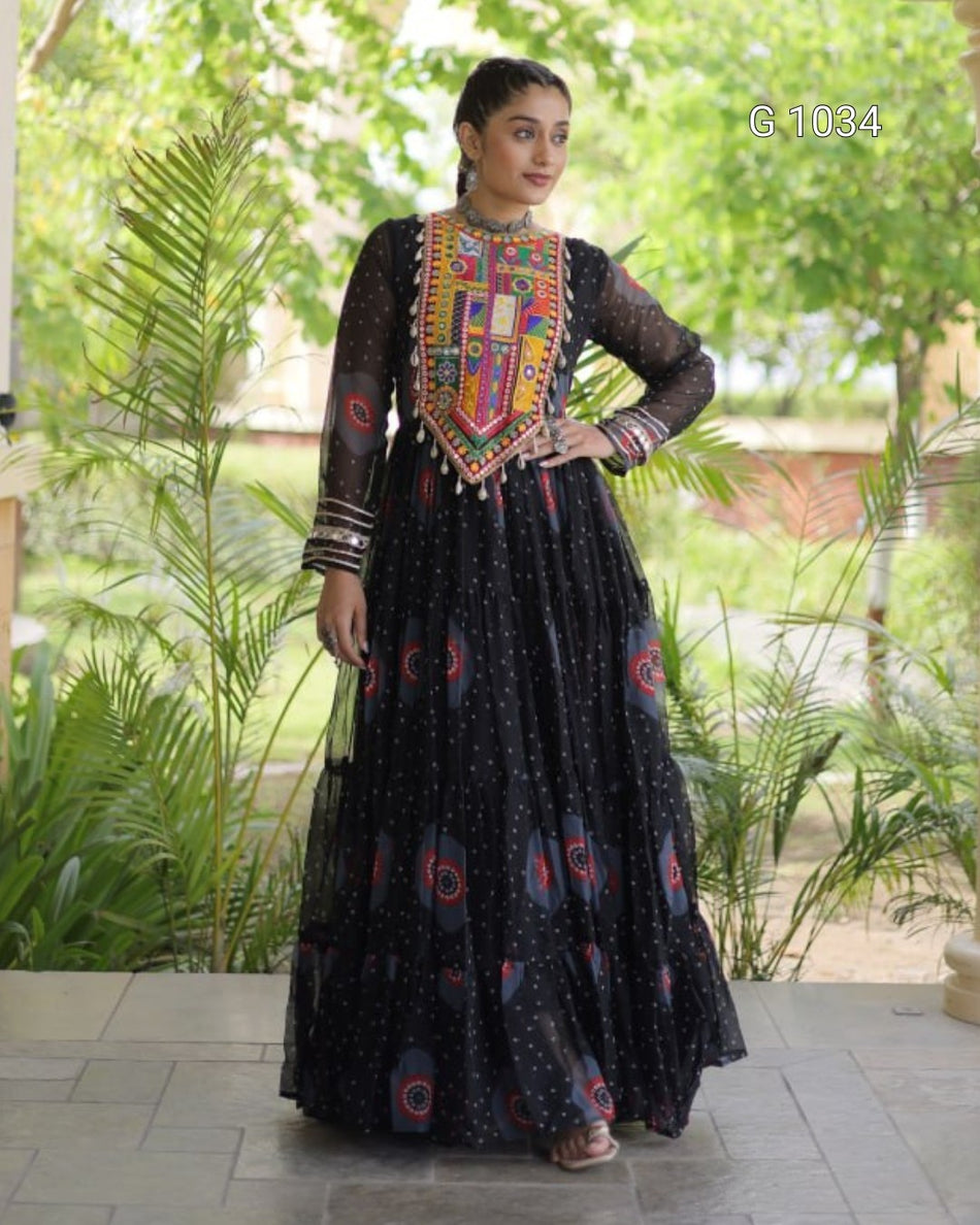 A woman modeling a black sheer Anarkali gown with a heavily embroidered multicolor neckline and scattered red and blue motifs on the skirt, set against a lush green outdoor background.