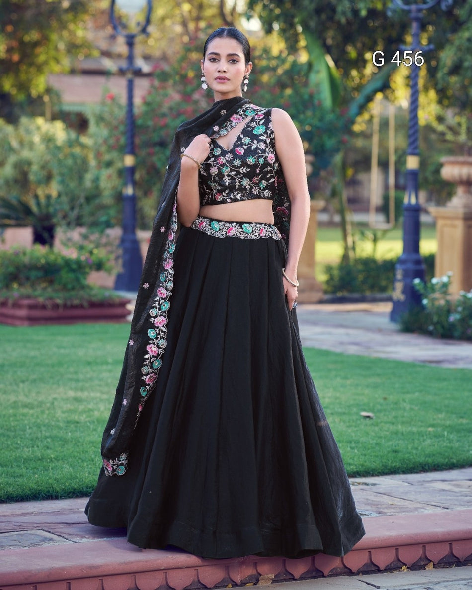 A woman stands outdoors on a paved walkway in a garden setting, wearing a black traditional outfit with floral embroidery. The outfit includes a sleeveless embroidered crop top, a long black skirt, and a matching embroidered dupatta.