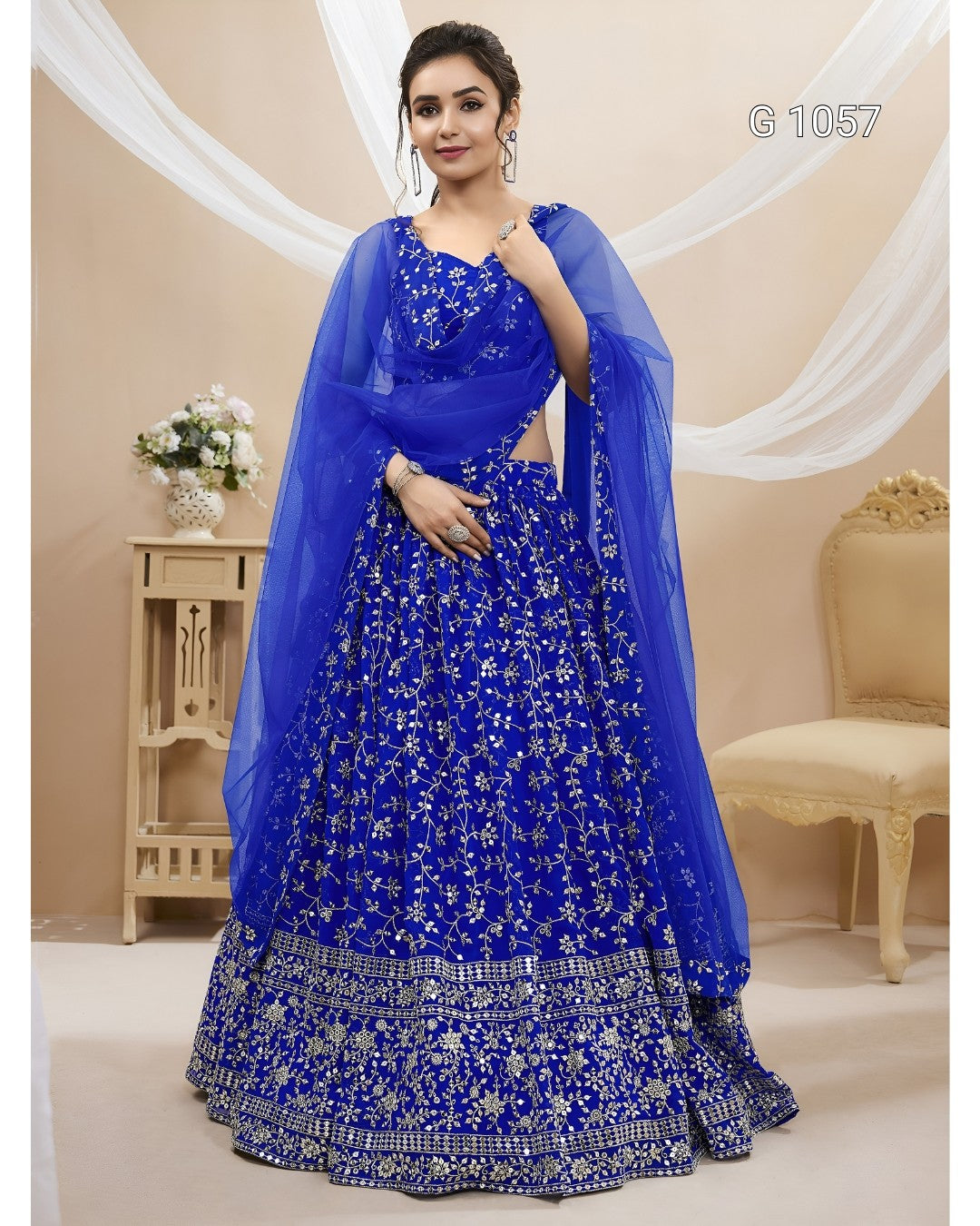 A woman models a royal blue heavily embroidered Indian ethnic gown with a matching sheer dupatta, set against a beige studio backdrop with draped white fabric and furniture.