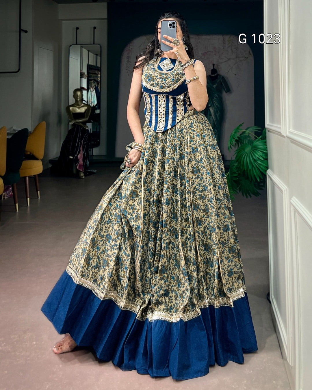 A woman takes a mirror selfie wearing a traditional Indian salwar suit with a heavily embroidered navy blue and gold skirt, a matching navy blue and gold embroidered bodice, and a solid navy blue flared hem. The background is a boutique interior.