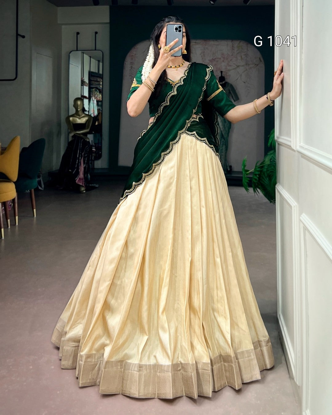 A woman takes a mirror selfie wearing a traditional Indian lehenga choli dress. The outfit features a voluminous cream-colored skirt (lehenga) with a metallic border, paired with a dark green sleeveless top (choli) and a matching draped scarf (saree drape) with gold embroidery.