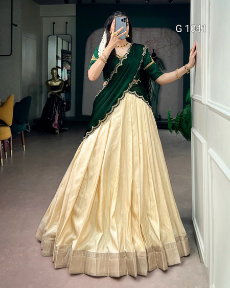 A woman takes a mirror selfie wearing a traditional Indian lehenga choli dress. The outfit features a voluminous cream-colored skirt (lehenga) with a metallic border, paired with a dark green sleeveless top (choli) and a matching draped scarf (saree drape) with gold embroidery.
