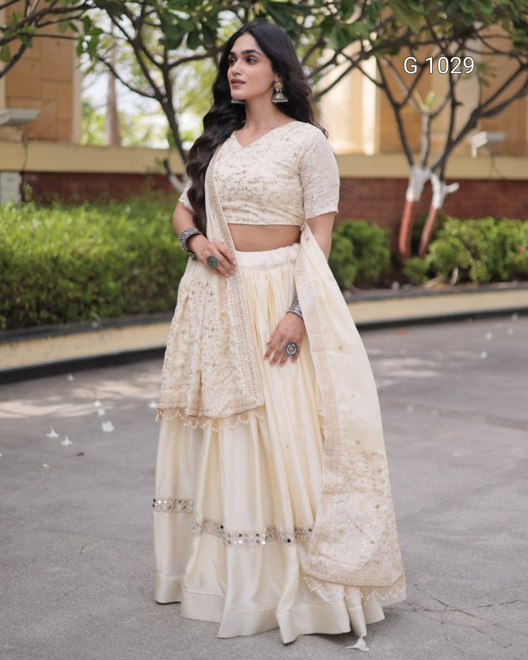 A woman modeling a cream-colored traditional Indian ethnic wear set consisting of a printed lace kurta, matching flared skirt, and draped dupatta, photographed outdoors on a paved walkway.