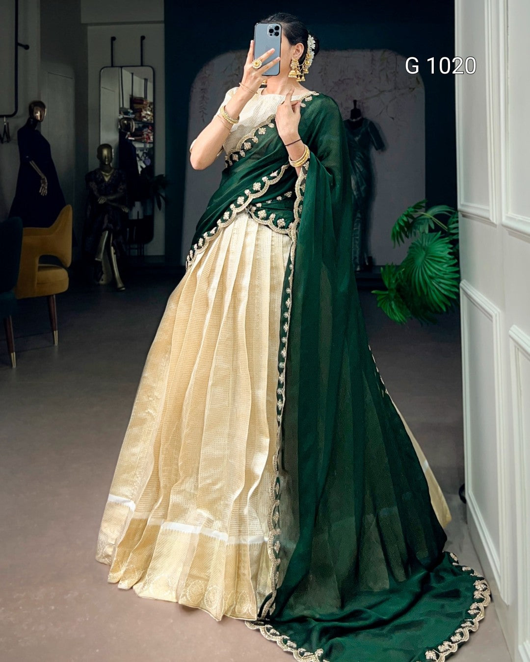 A woman takes a mirror selfie in a boutique wearing a traditional Indian outfit consisting of a cream-colored flared skirt (lehenga), a dark green draped fabric (sharara/pallu), and a matching sheer green dupatta with gold embroidered borders. The background shows store fixtures, mannequins, and a plant.