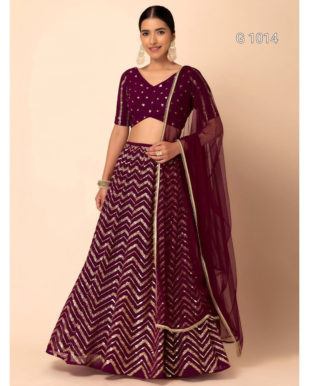A woman modeling a deep purple lehenga choli set with gold chevron patterns and a matching sheer dupatta, set against a solid beige background.