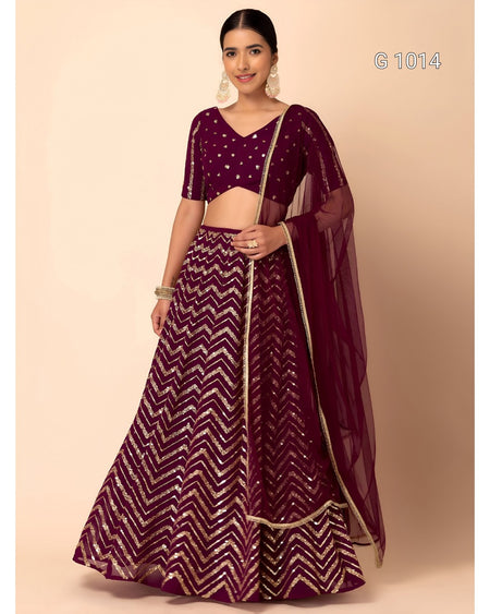 A woman modeling a deep purple lehenga choli set with gold chevron patterns and a matching sheer dupatta, set against a solid beige background.