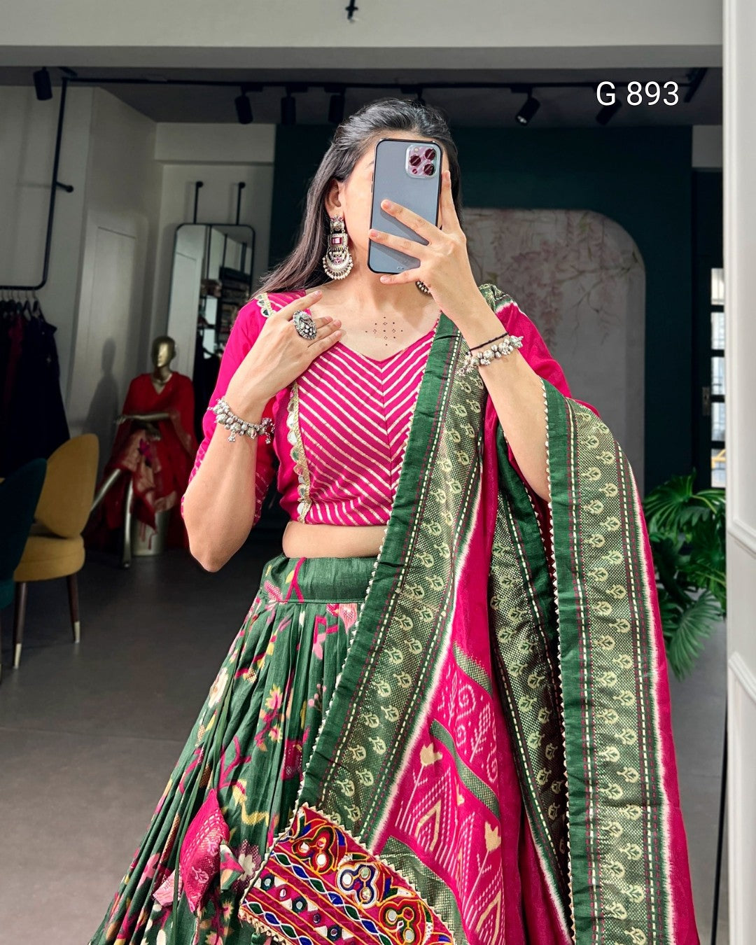 Woman taking a mirror selfie wearing a pink and green traditional outfit with a blurred background.