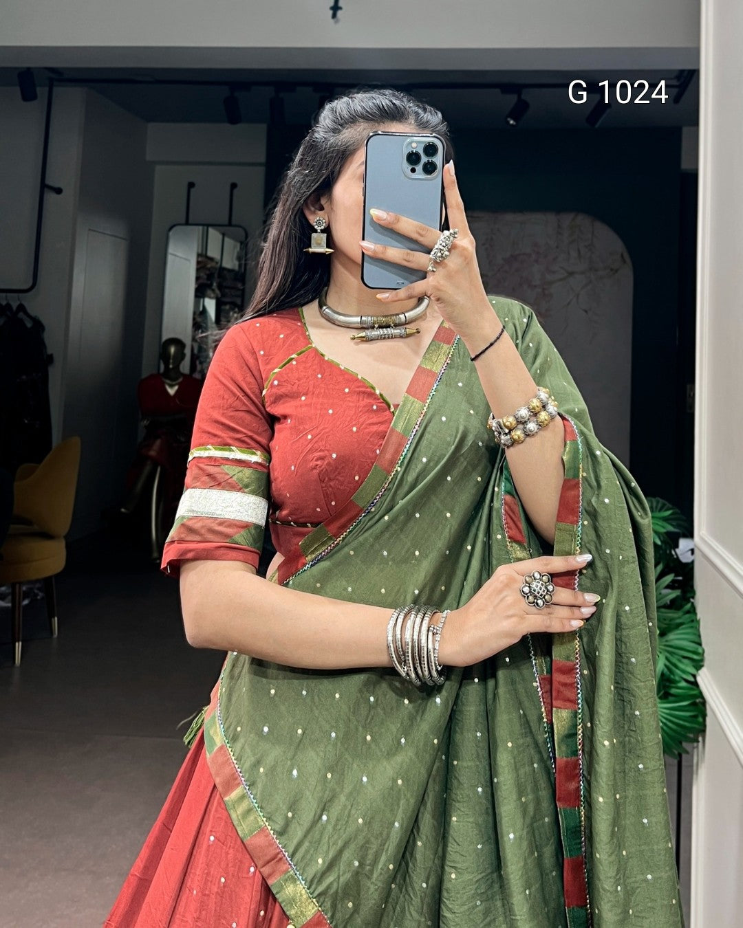 A woman takes a mirror selfie wearing a traditional Indian outfit consisting of a red blouse with green and white stripes, and a sage green saree with small white motifs and red borders. She is wearing silver jewelry and holding a blue smartphone.