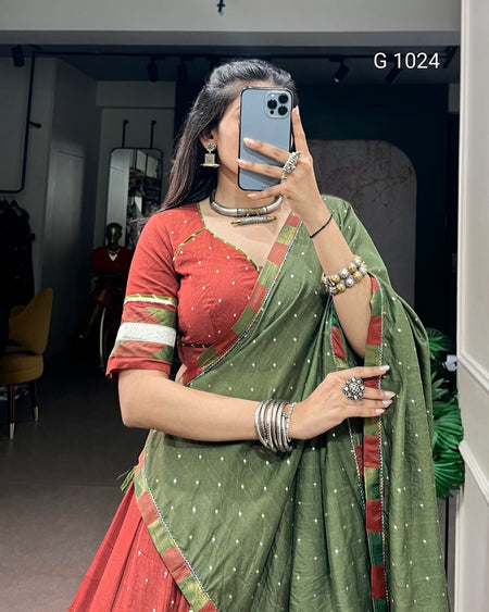 A woman takes a mirror selfie wearing a traditional Indian outfit consisting of a red blouse with green and white stripes, and a sage green saree with small white motifs and red borders. She is wearing silver jewelry and holding a blue smartphone.