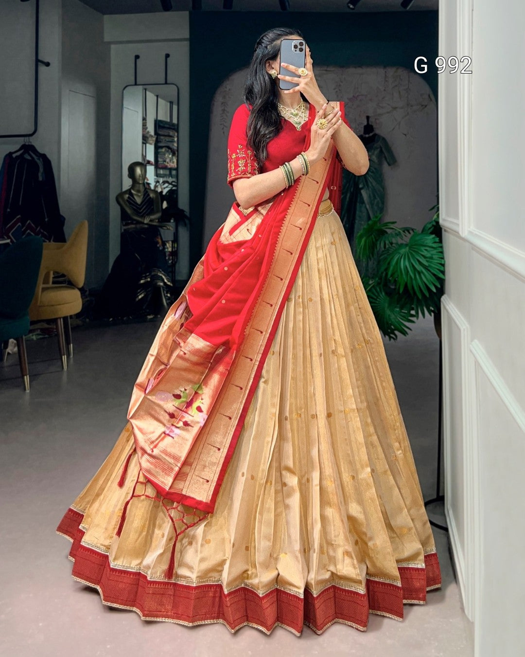 A woman takes a mirror selfie wearing a traditional Indian outfit consisting of a beige flared lehenga skirt with a red border, a red blouse, and a red dupatta with gold zari detailing.