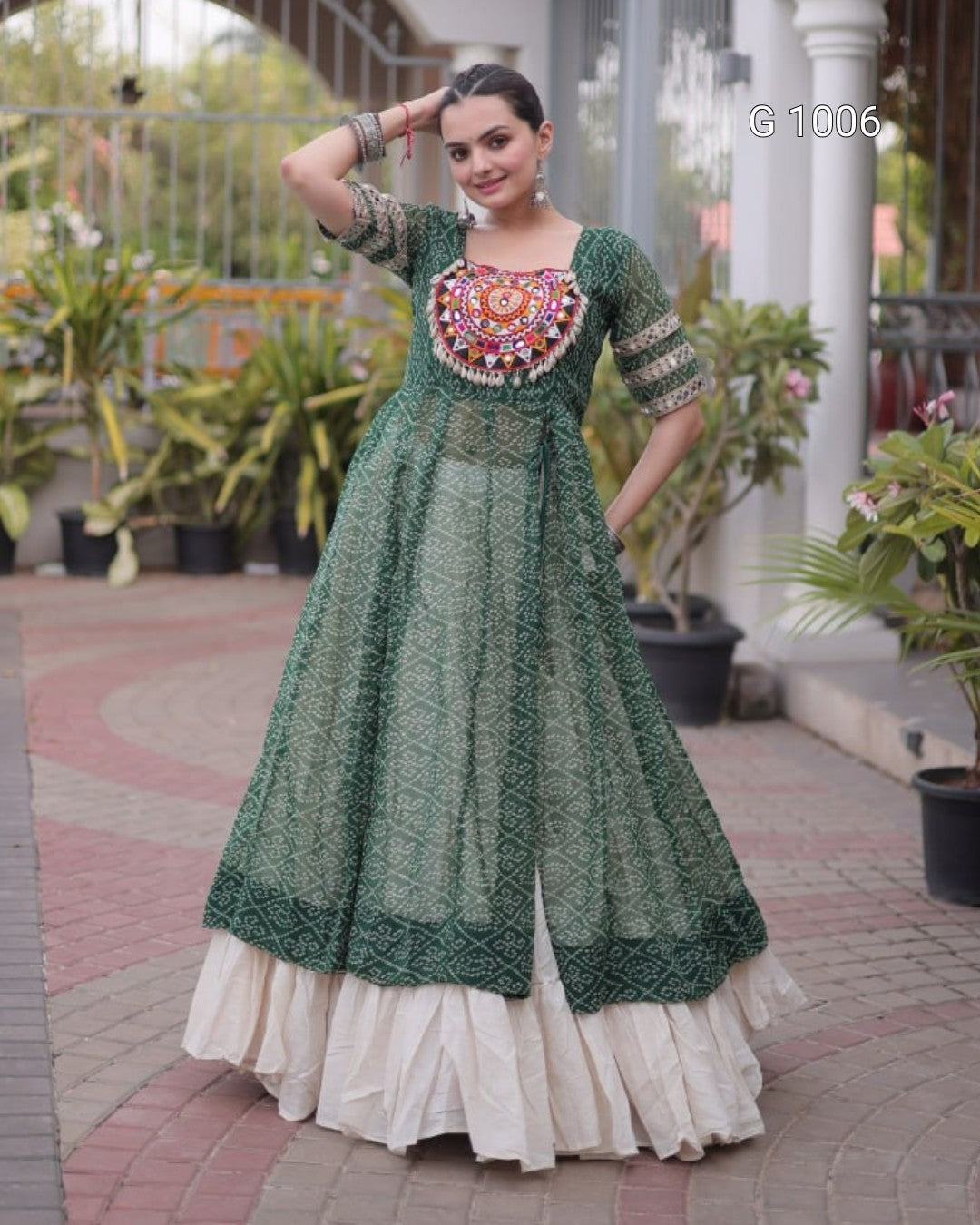 A woman modeling a traditional Indian Anarkali gown in forest green with intricate multicolor embroidery on the chest and a layered cream-colored flared skirt, set against an outdoor patio background with potted plants.