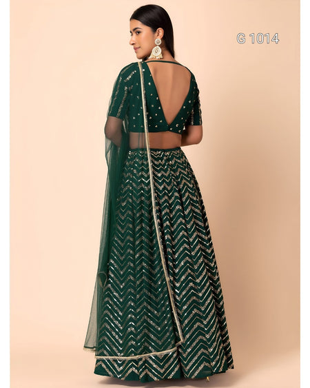 A woman modeling a dark green traditional outfit with a chevron patterned skirt, matching blouse, and sheer dupatta, shown from the back against a solid beige background.
