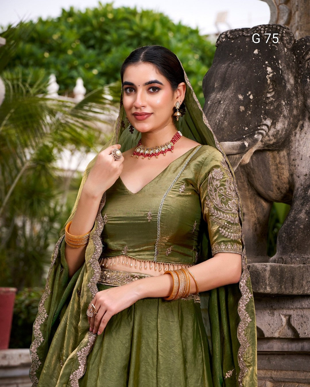 A woman wearing an olive green embroidered traditional outfit with a matching dupatta, standing outdoors beside a stone elephant statue. The outfit has gold-toned embroidery and a metallic embellished waist detail.