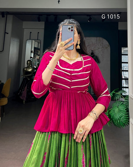 A woman takes a mirror selfie wearing a vibrant magenta and green traditional outfit with gold jewelry. The top is bright magenta with horizontal cream stripes, and the skirt is green with pink vertical stripes.