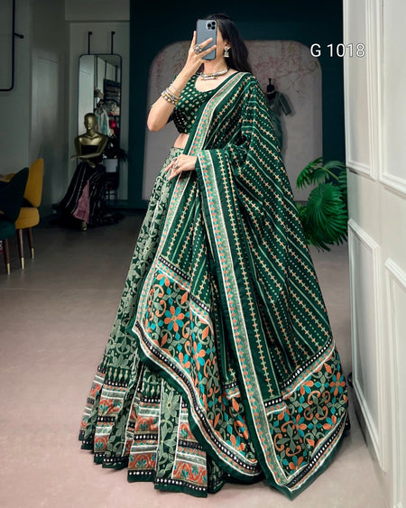 A woman takes a mirror selfie wearing a dark green traditional Indian lehenga choli set with intricate multicolor embroidery and geometric patterns. The setting is an indoor boutique or room with a mannequin visible in the background.