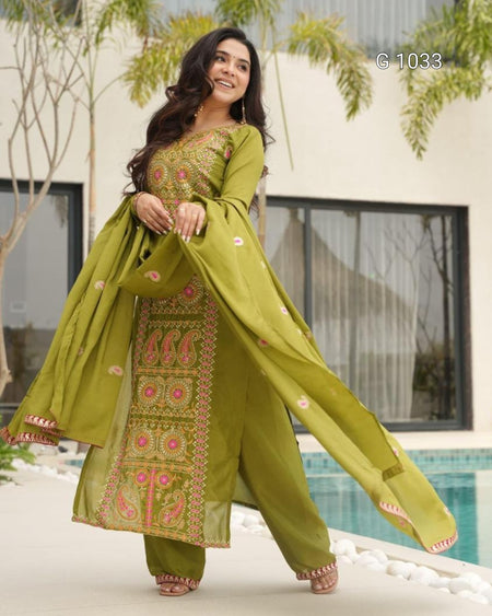 A woman models a three-piece olive green ethnic salwar kameez suit with intricate pink and gold embroidery, standing outdoors near a pool.